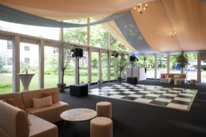 The interior of a modern event marquee featuring a black and white checkered dance floor, lounge seating with peach-colored pillows, and large glass windows overlooking a green garden.
