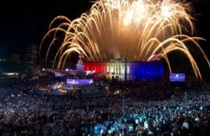 A night view of a large outdoor concert stage in front of a palace illuminated in red, white, and blue, with massive golden fireworks exploding overhead