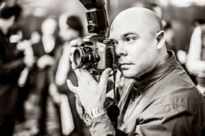 A black and white, close up profile shot of a man looking through the viewfinder of a professional DSLR camera. He is wearing a shirt and a watch. The background shows a blurred crowd of people.