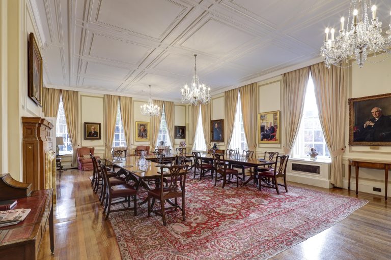 A wide view of a grand conference room featuring two long wooden tables with red upholstered chairs, crystal chandeliers, and oil portraits on the walls.