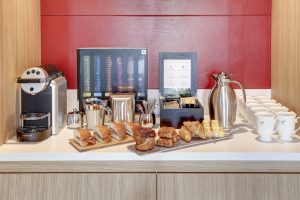 Close up of office refreshment station with Nespresso machine, sandwiches, pastries, tea, and coffee cups against red wall.