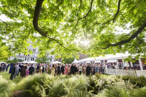 Guests mingling at a large garden party, gathered in front of a marquee with sunlight shining through the tree canopy and historic buildings in the background.