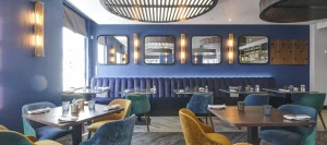 Pegasus Bar dining room at Inner Temple with deep blue banquette seating, square wooden tables, and mixed teal, blue, and mustard velvet chairs beneath large slatted ceiling lights and wall mirrors.
