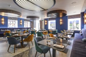 Another angle of Pegasus Bar’s interior, highlighting the contemporary ceiling feature, large windows, and neatly set tables with a mix of soft blue and yellow seating.