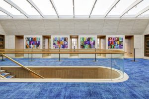 Expansive Inner Temple hallway with blue carpet, glass balustrade staircase, colourful portrait wall art, and bookshelves.