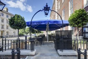 Outdoor terrace of Pegasus Bar at Inner Temple, with blue canopy, black tables and chairs, and surrounding potted trees on a sunny day.