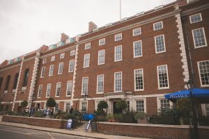 A wide angle photograph of a grand Georgian style red brick building with white stone accents, tall sash windows, and decorative cornices under a cloudy sky. The multi story structure features arched doorways, pediments, manicured shrubs, and potted plants in front. People stand on the pavement nearby, with a prominent blue 'Pegasus' umbrella and signage visible at the entrance on a paved street.