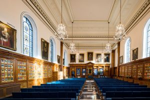 Grand hall in the Inner Temple with high white ceiling and multiple sparkling chandeliers, light wood panelled walls displaying full length portraits and wooden cabinets with shields, tall arched stained glass windows allowing warm sunlight, and rows of blue upholstered chairs facing a central wooden dais on a checkered marble floor.