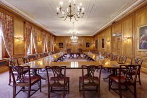 Long, elegant wood panelled room in the Inner Temple with polished oak walls and ceiling, pink floral curtains on tall windows, multiple crystal chandeliers, wall sconces, portraits of figures in ornate frames, and a large oval polished wood conference table surrounded by red upholstered chairs on red carpet.