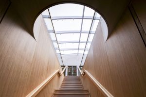 Wood panelled corridor with curved walls leading to wooden stairs flanked by railings, under a large circular glass skylight allowing natural light.
