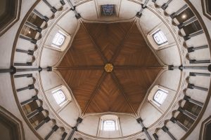 Fish eye upward view of Temple Church's octagonal wooden ceiling with dark beams forming geometric patterns around a central golden ornament, supported by light stone arches and columns, with small stained glass windows and white wall panels.