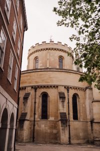 Exterior view of Temple Church's circular stone tower in warm beige with narrow arched windows and buttresses, topped by a crenellated parapet and cross, beside a red brick building, framed by green tree branches against a pale sky.
