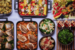 Close-up of an elegant row of assorted canapés arranged in lines on a light marble surface, including smoked salmon bites, breaded fried balls with red sauce, vegetable-topped crisps, and grilled meat pieces garnished with herbs and edible flowers.