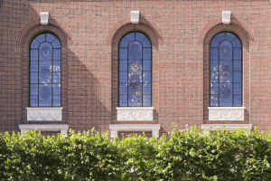 Three tall arched stained glass windows with purple, blue, and multicolored panels in a red brick wall with white stone sills and decorative cornices, framed by green hedges below.